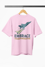Pink t-shirt with a graphic of a jet and 'Embrace Transformation' text on a hanger against a white background.