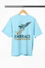 Light blue t-shirt with airplane graphic and 'Embrace Transformation' text on a hanger.