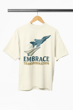 Beige t-shirt with airplane graphic and 'Embrace Transformation' text on a hanger.