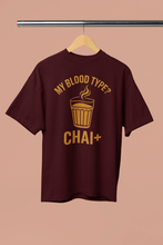 Maroon t-shirt with yellow text 'My Blood Type? CHAI+' on a hanger against a beige background