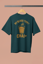 Green t-shirt with yellow text 'My blood type? CHAI+' on a hanger against a beige background