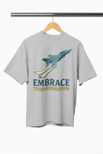 Gray t-shirt with airplane graphic and 'Embrace Transformation' text on a hanger against a white background